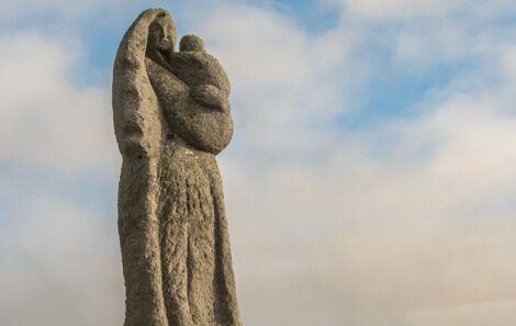 A stone statue depicts a veiled woman holding a child against a backdrop of a cloudy sky.