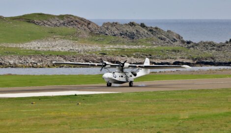 A white seaplane with two propellers lands on a paved airstrip near a rocky coastline with grassy fields and a body of water in the background.