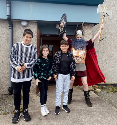 Three children stand in front of a building entrance, smiling. Behind them, a person dressed in a Viking costume with a shield and axe poses enthusiastically.