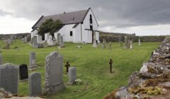 A white, stone church stands amidst a grassy cemetery with numerous tombstones, under a cloudy sky.