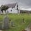 A white, stone church stands amidst a grassy cemetery with numerous tombstones, under a cloudy sky.