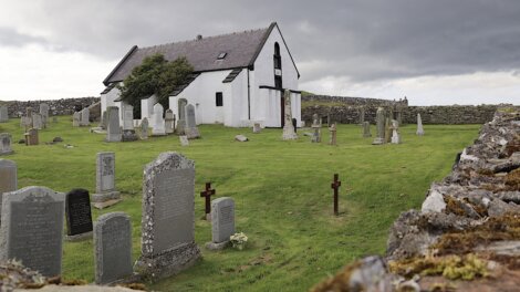 A white, stone church stands amidst a grassy cemetery with numerous tombstones, under a cloudy sky.