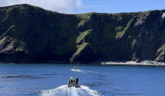 Two small boats with people in high-visibility jackets navigate a calm body of water near a rugged, grassy cliff coastline under a clear blue sky.