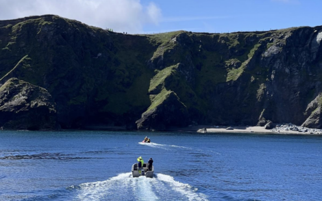 Two small boats with people in high-visibility jackets navigate a calm body of water near a rugged, grassy cliff coastline under a clear blue sky.