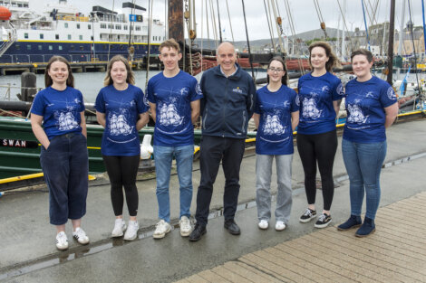 A group of seven people wearing blue shirts and jeans pose for a photo in front of boats at a harbor.