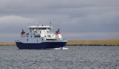 A large blue and white cargo ship sails on a body of water, with grasslands and cloudy skies in the background.