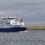 A large blue and white cargo ship sails on a body of water, with grasslands and cloudy skies in the background.
