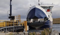 A ferry with its loading ramp lowered docking at a port. The front of the ferry is open, revealing the interior vehicle deck. The port area has yellow railings and a docking structure.