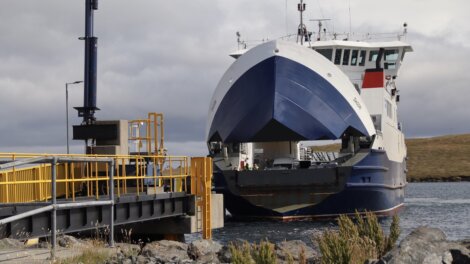 A ferry with its loading ramp lowered docking at a port. The front of the ferry is open, revealing the interior vehicle deck. The port area has yellow railings and a docking structure.