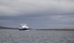 A ferry sails on calm waters under a cloudy sky, approaching a low, grassy coastline in the distance.