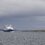 A ferry sails on calm waters under a cloudy sky, approaching a low, grassy coastline in the distance.