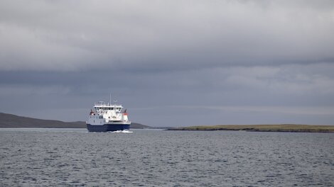 A ferry sails on calm waters under a cloudy sky, approaching a low, grassy coastline in the distance.