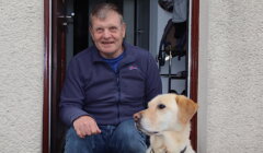 A person sits in a doorway next to a Labrador retriever. The person is wearing a blue jacket and jeans, and the dog is wearing a harness.