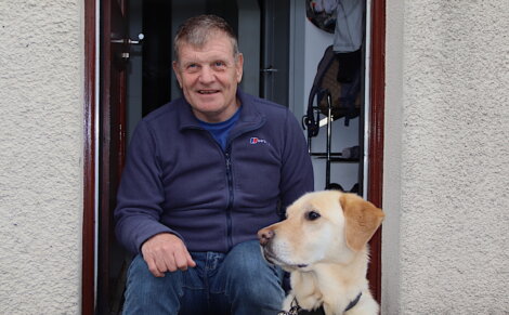 A person sits in a doorway next to a Labrador retriever. The person is wearing a blue jacket and jeans, and the dog is wearing a harness.