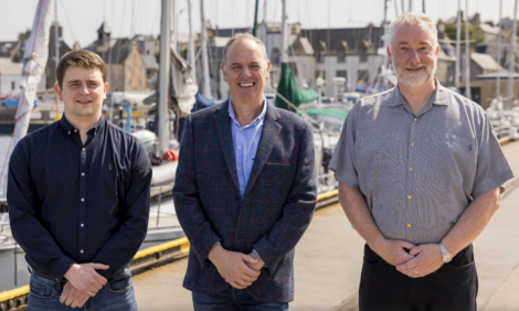 Three men standing outdoors in front of a marina with boats and buildings in the background, all smiling and facing the camera.