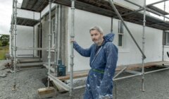 A man in a blue jumpsuit, covered in white paint splatters, stands on a gravel surface holding a scaffold pole beside a white house under construction.