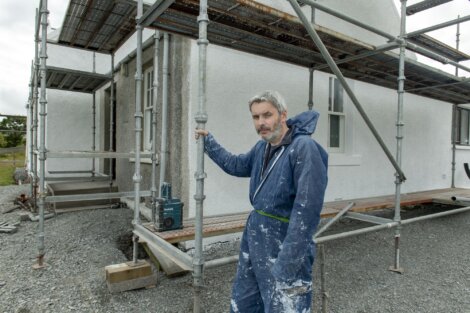 A man in a blue jumpsuit, covered in white paint splatters, stands on a gravel surface holding a scaffold pole beside a white house under construction.