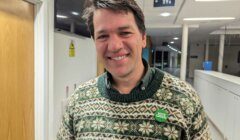 A man wearing a patterned sweater and a "Vote Green" button smiles in a brightly lit indoor hallway.