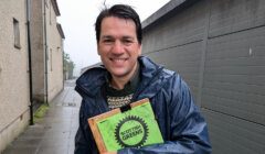 A person in a raincoat holds a clipboard with a "Scottish Greens" logo while standing in a wet, narrow alleyway.