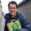 A person in a raincoat holds a clipboard with a "Scottish Greens" logo while standing in a wet, narrow alleyway.