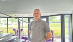 A man in a gray polo shirt stands by a staircase in a modern building with large windows and purple chairs.