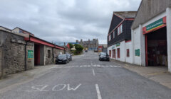 A narrow, uphill street with "SLOW" painted on the road, flanked by brick buildings on both sides. Two cars are parked on the street. Cloudy sky in the background.