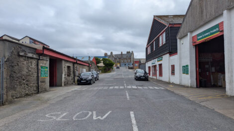 A narrow, uphill street with "SLOW" painted on the road, flanked by brick buildings on both sides. Two cars are parked on the street. Cloudy sky in the background.