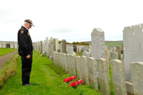 A man in uniform stands solemnly in a cemetery, facing headstones with red wreaths placed at their bases.