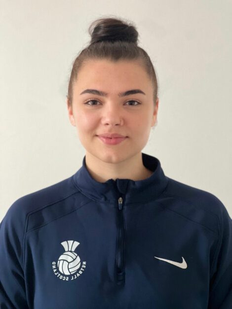 Person wearing a dark blue sports jacket with a netball emblem and a Nike logo, hair tied in a bun, smiling.