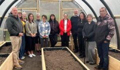 A group of people stands inside a greenhouse structure with raised garden beds filled with soil. They seem to be posing for the photo.