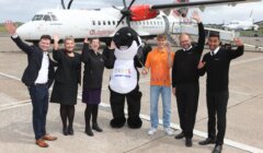 A group of six people, including one in an orca costume, stand in front of a Loganair plane, all raising their hands and smiling.