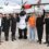 A group of six people, including one in an orca costume, stand in front of a Loganair plane, all raising their hands and smiling.