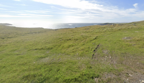 A grassy field stretches toward a distant view of the sea under a partly cloudy sky, with sunlight reflecting off the water.