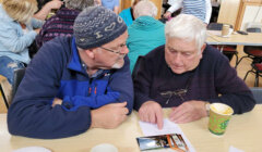 Two older men sit at a table, discussing or reading a document. One man points to the document. There are other people sitting and conversing in the background.