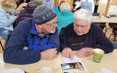 Two older men sit at a table, discussing or reading a document. One man points to the document. There are other people sitting and conversing in the background.