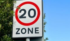 A road sign with a red circle and the number "20" indicating a 20 mph speed limit zone, positioned next to green foliage against a clear blue sky.