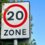 A road sign with a red circle and the number "20" indicating a 20 mph speed limit zone, positioned next to green foliage against a clear blue sky.