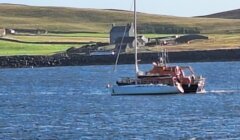 A sailboat and a small motorized boat are on the water near a distant shore with green fields and buildings.