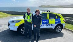 Two police officers stand in front of a police car near a coastal area with the ocean and hills in the background.