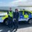 Two police officers stand in front of a police car near a coastal area with the ocean and hills in the background.