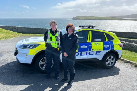 Two police officers stand in front of a police car near a coastal area with the ocean and hills in the background.