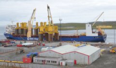 A cargo ship is docked at a port, loaded with large yellow structures and featuring cranes. Buildings and equipment are seen in the foreground, with a body of water and land in the background.