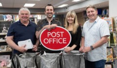 Four people standing indoors holding a red "Post Office" sign, with three mail bags in front of them and shelves of items in the background.