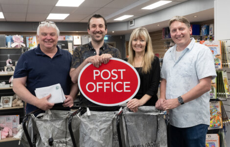 Four people standing indoors holding a red "Post Office" sign, with three mail bags in front of them and shelves of items in the background.