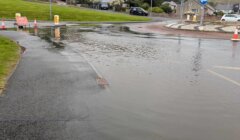 Flooded roundabout with traffic cones and a road closed sign indicating the area is impassable due to heavy rain. Surrounding residential houses and lawns are visible in the background.