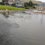 Flooded roundabout with traffic cones and a road closed sign indicating the area is impassable due to heavy rain. Surrounding residential houses and lawns are visible in the background.