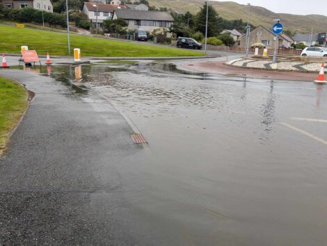 Flooded roundabout with traffic cones and a road closed sign indicating the area is impassable due to heavy rain. Surrounding residential houses and lawns are visible in the background.