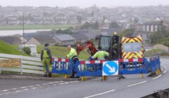 Workers in high-visibility clothing operate an excavator and perform roadwork behind barriers on a street. Buildings and green lawns are visible in the background on a cloudy day.