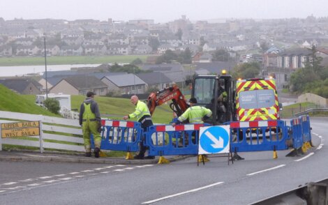 Workers in high-visibility clothing operate an excavator and perform roadwork behind barriers on a street. Buildings and green lawns are visible in the background on a cloudy day.