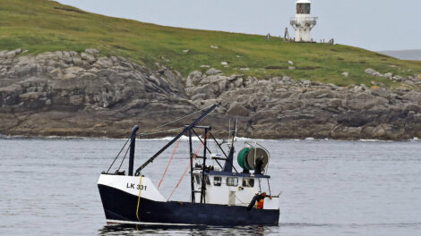 A small fishing boat with the designation LK 331 sails near a rocky shoreline with a white lighthouse in the background.